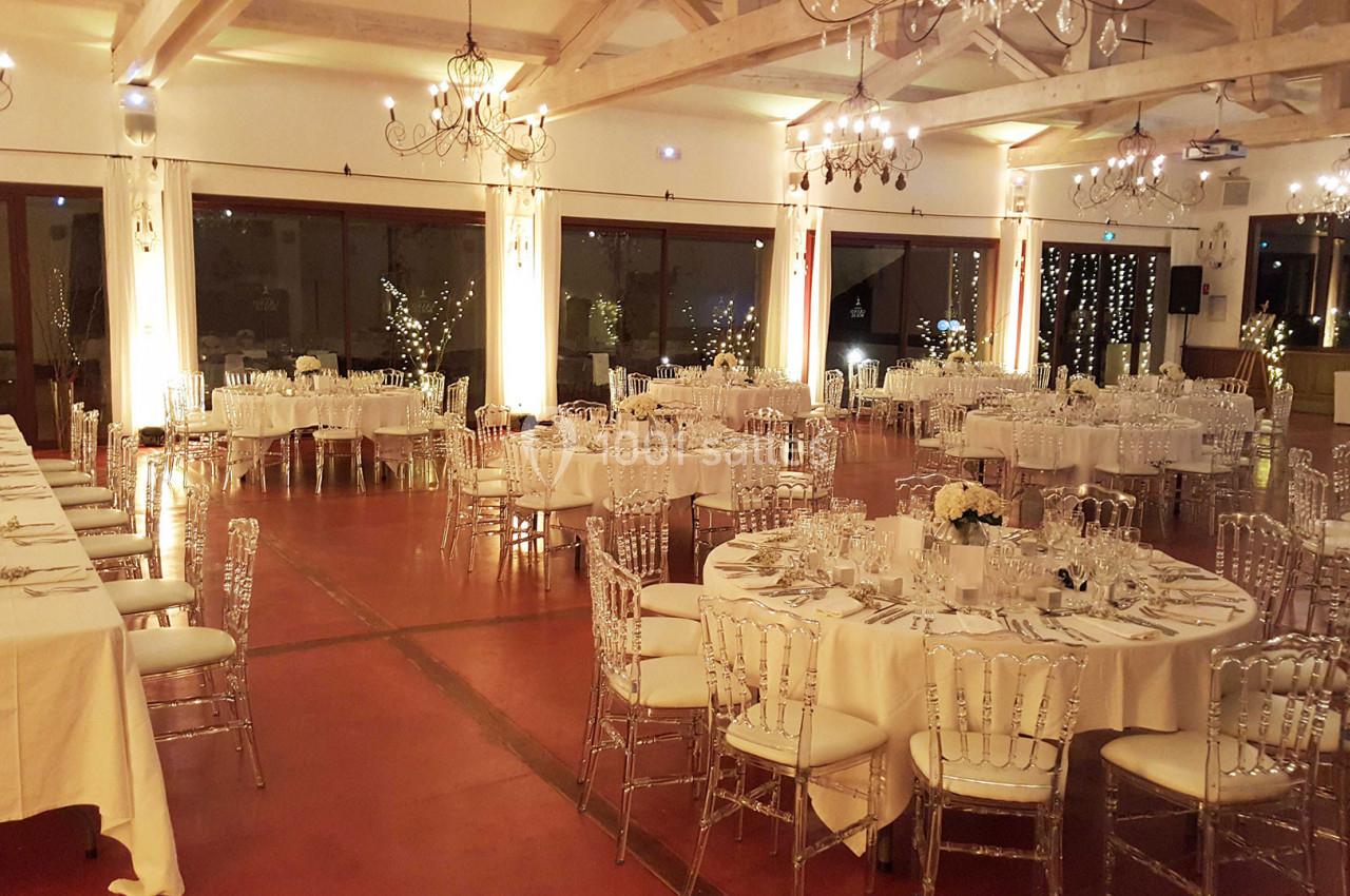 Salle de réception élégamment décorée avec des tables rondes dressées, des chaises transparentes et des lumières tamisées.