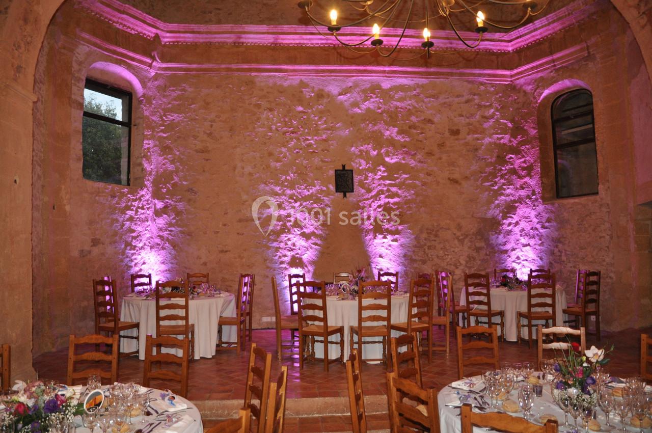 Salle en pierre avec tables dressées pour un événement, éclairée par des lumières roses et un lustre au plafond.