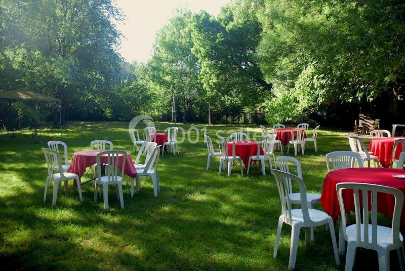 Chaises blanches et tables avec nappes rouges disposées sur une pelouse entourée d'arbres sous un ciel dégagé.