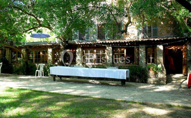 Façade d'une maison en pierre entourée d'arbres, avec une longue table dressée sur une terrasse ombragée.