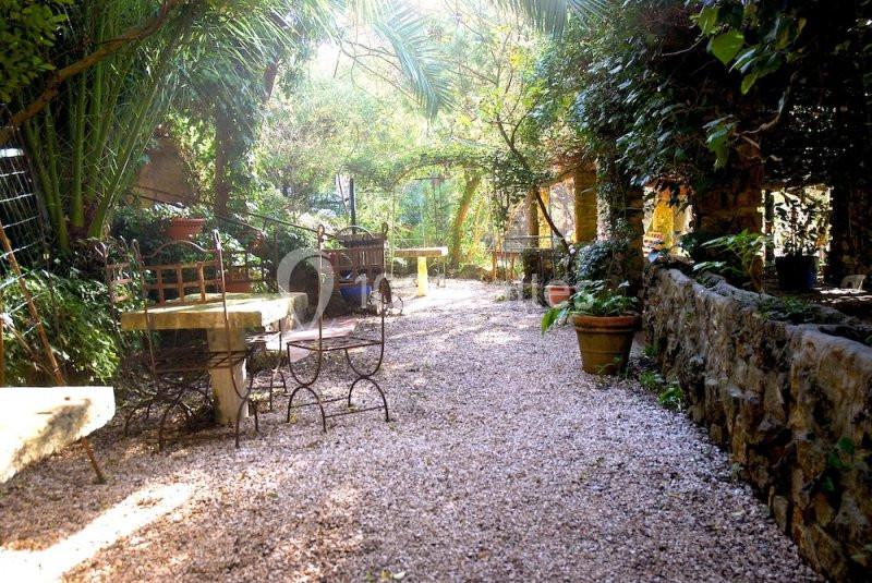 Allée de gravier bordée de végétation luxuriante, avec des tables et chaises en fer forgé dans un jardin ombragé.