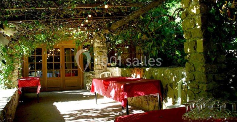 Terrasse ombragée avec tables recouvertes de nappes rouges, entourée de végétation et éclairée par des guirlandes lumineuses.
