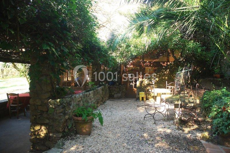 Cour extérieure ombragée avec des tables, des chaises en métal et des plantes grimpantes entourant un bâtiment en pierre.