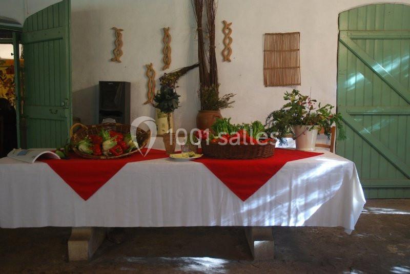 Table décorée avec une nappe blanche et rouge, garnie de paniers de légumes frais, dans une pièce lumineuse aux volets verts.
