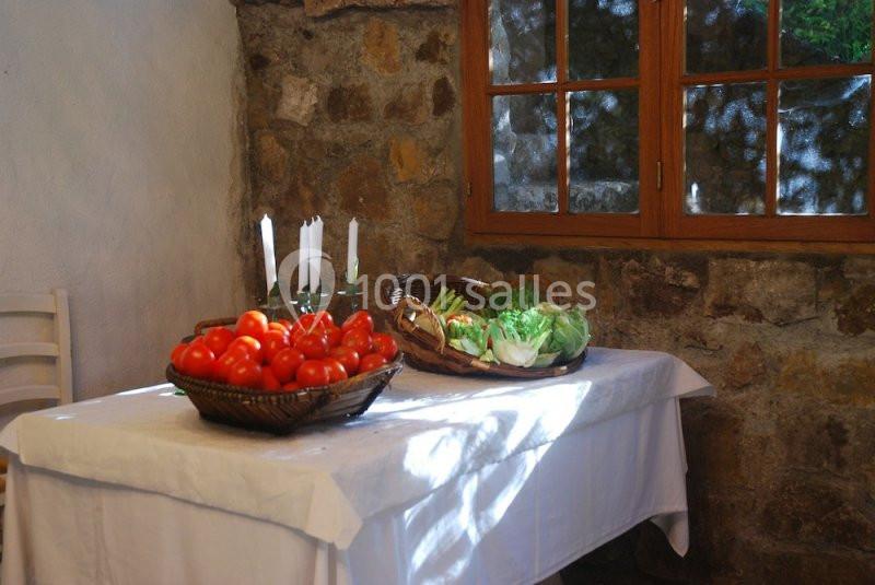 Des paniers de tomates et de légumes verts posés sur une table blanche près d'une fenêtre dans une pièce en pierre.