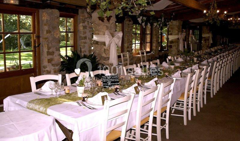 Salle de réception décorée avec de longues tables dressées, nappes blanches et éléments végétaux, près de fenêtres donnant…