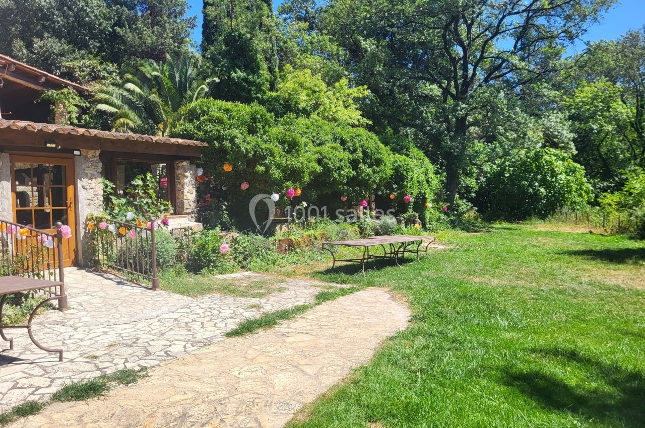 Jardin verdoyant avec terrasse en pierre, tables en métal et guirlandes colorées suspendues près d'une maison en pierre.