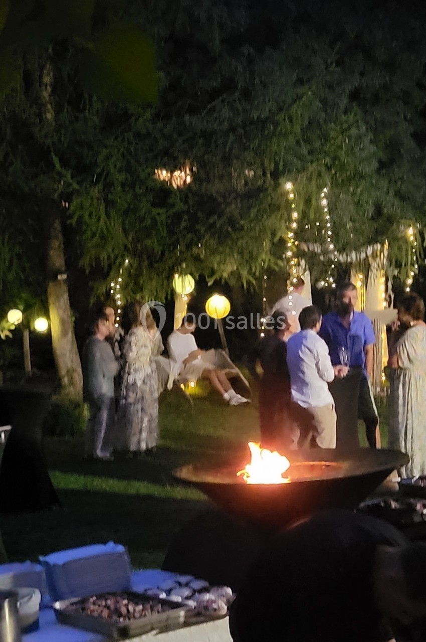 Groupe de personnes discutant près d'un brasero dans un jardin éclairé par des guirlandes lumineuses et des lanternes.