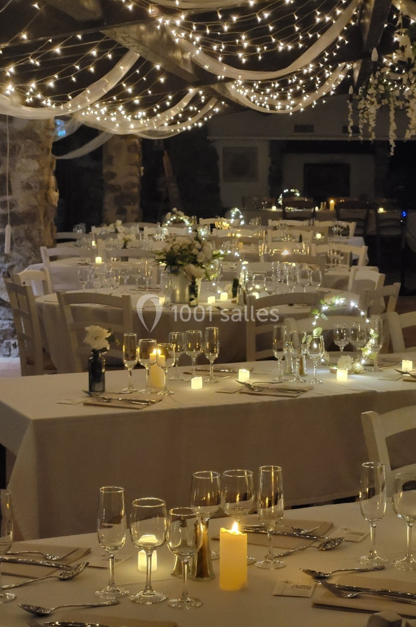 Salle de réception décorée avec des guirlandes lumineuses, des tables dressées avec nappes blanches et des bougies allumées.