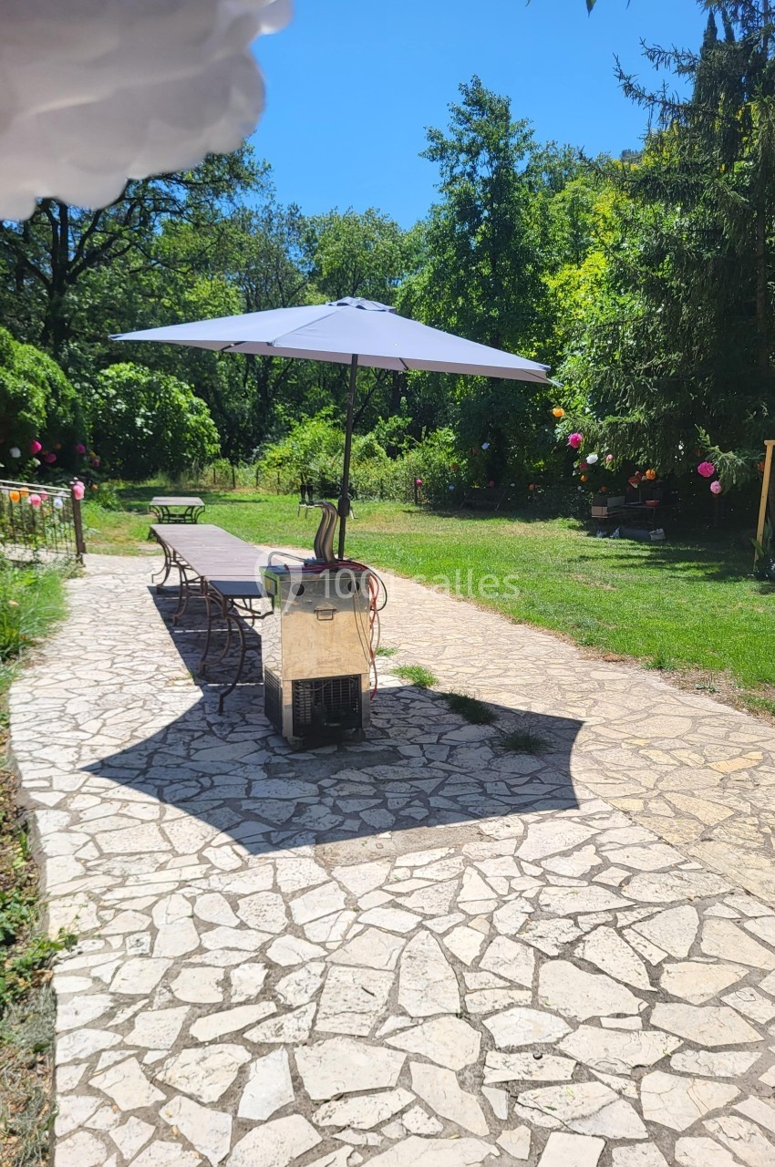 Allée en pierre bordée de verdure avec une table longue, des chaises et un parasol sous un ciel ensoleillé.