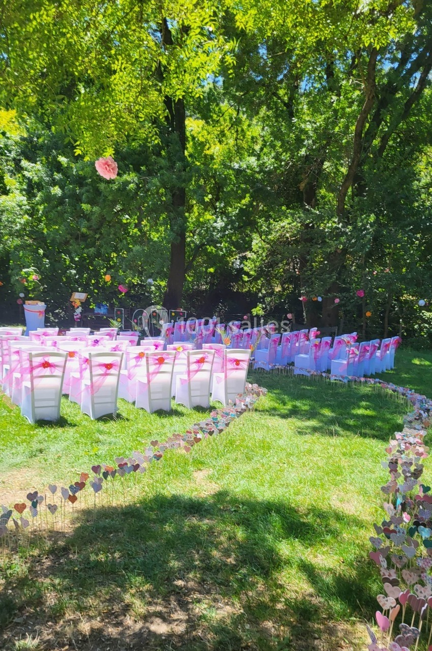 Chaises blanches décorées de rubans roses alignées en extérieur, entourées d'arbres et de décorations florales.