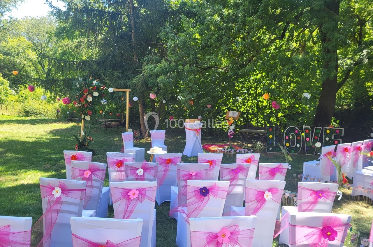 Chaises blanches décorées de rubans roses disposées en extérieur pour une cérémonie, entourées de verdure et de fleurs.