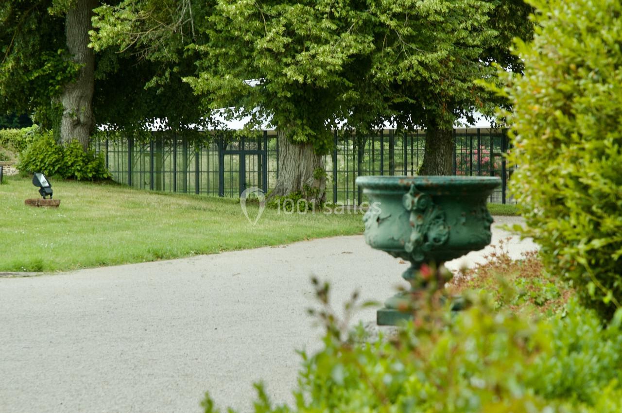 Allée bordée de verdure avec une grande vasque décorative au premier plan et une serre en arrière-plan.