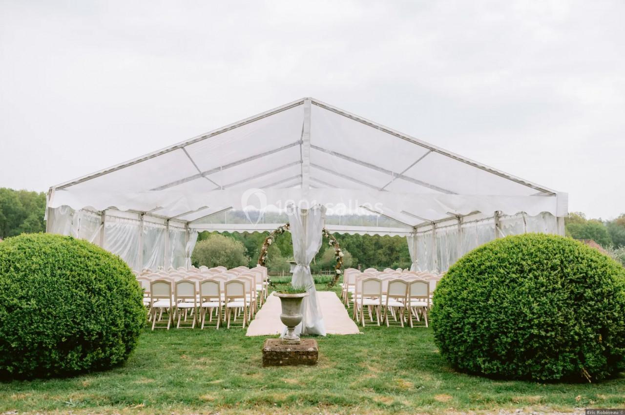Tente blanche transparente installée en extérieur, avec des rangées de chaises et une arche décorée pour une cérémonie.