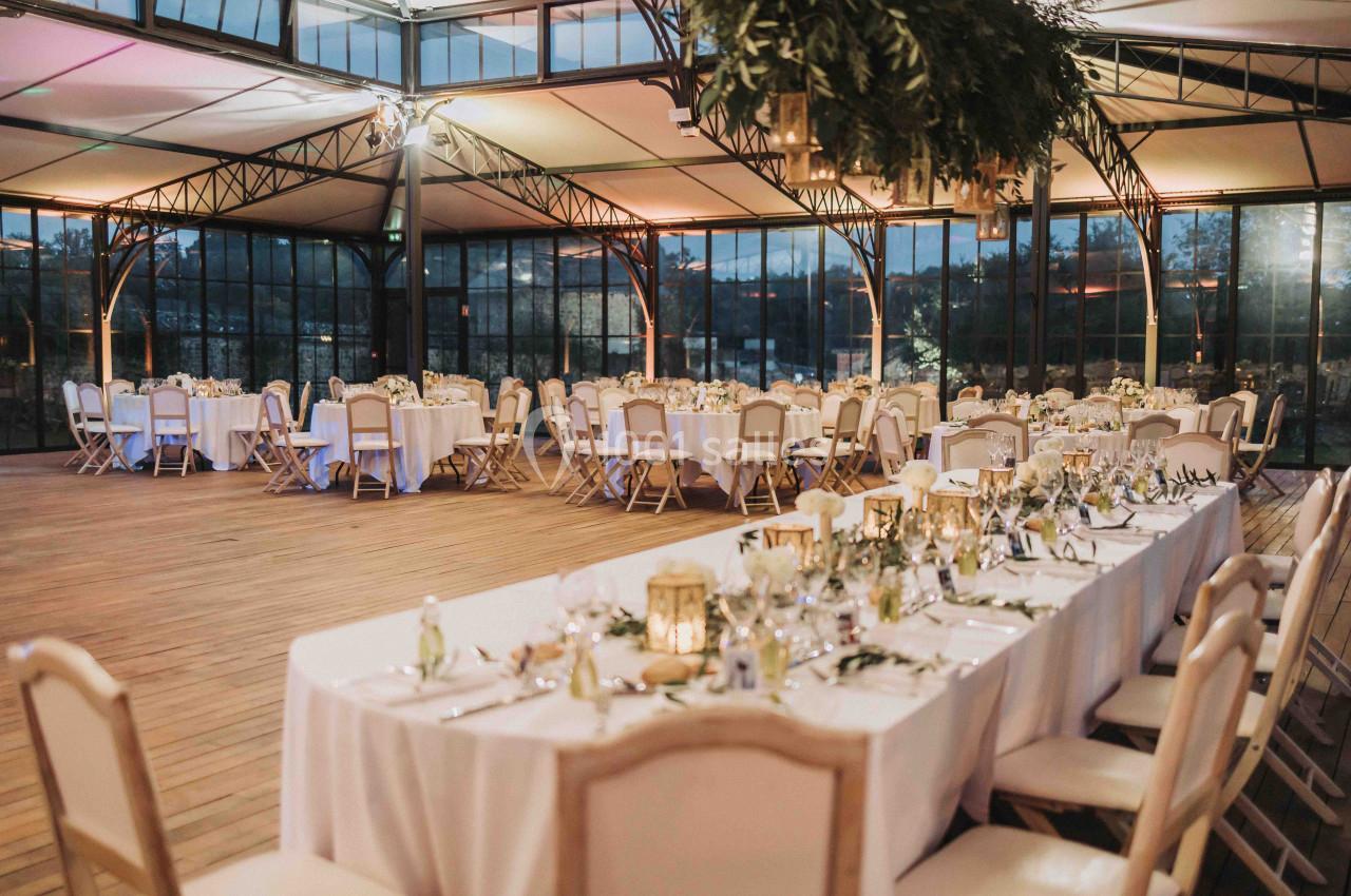 Salle de réception lumineuse avec tables décorées pour un événement, entourée de grandes baies vitrées et d'une structure…