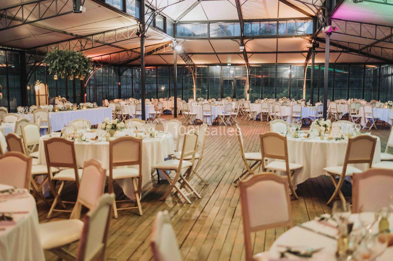 Salle de réception spacieuse avec tables rondes décorées de fleurs, chaises alignées et plafond en verrière.