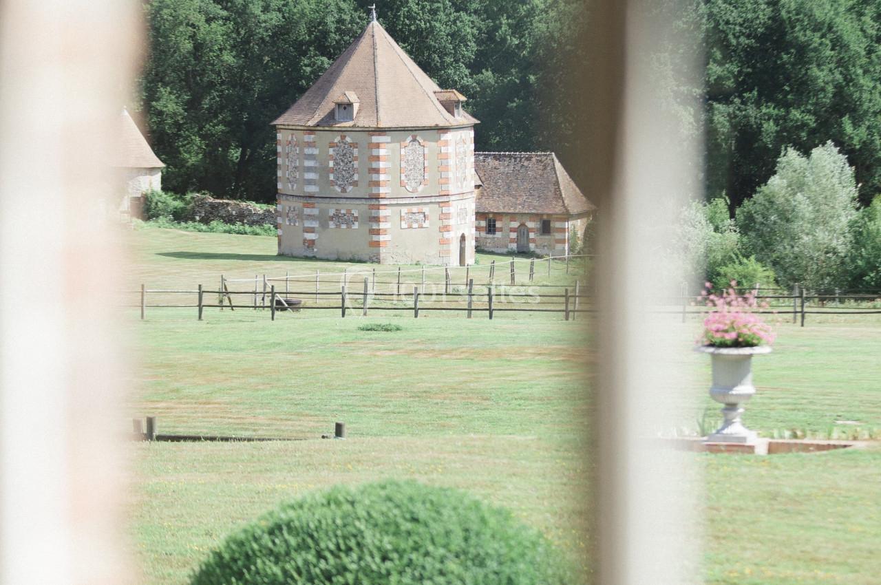 Vue d'un jardin verdoyant avec un bâtiment à colombages en arrière-plan, encadré par une fenêtre floue au premier plan.