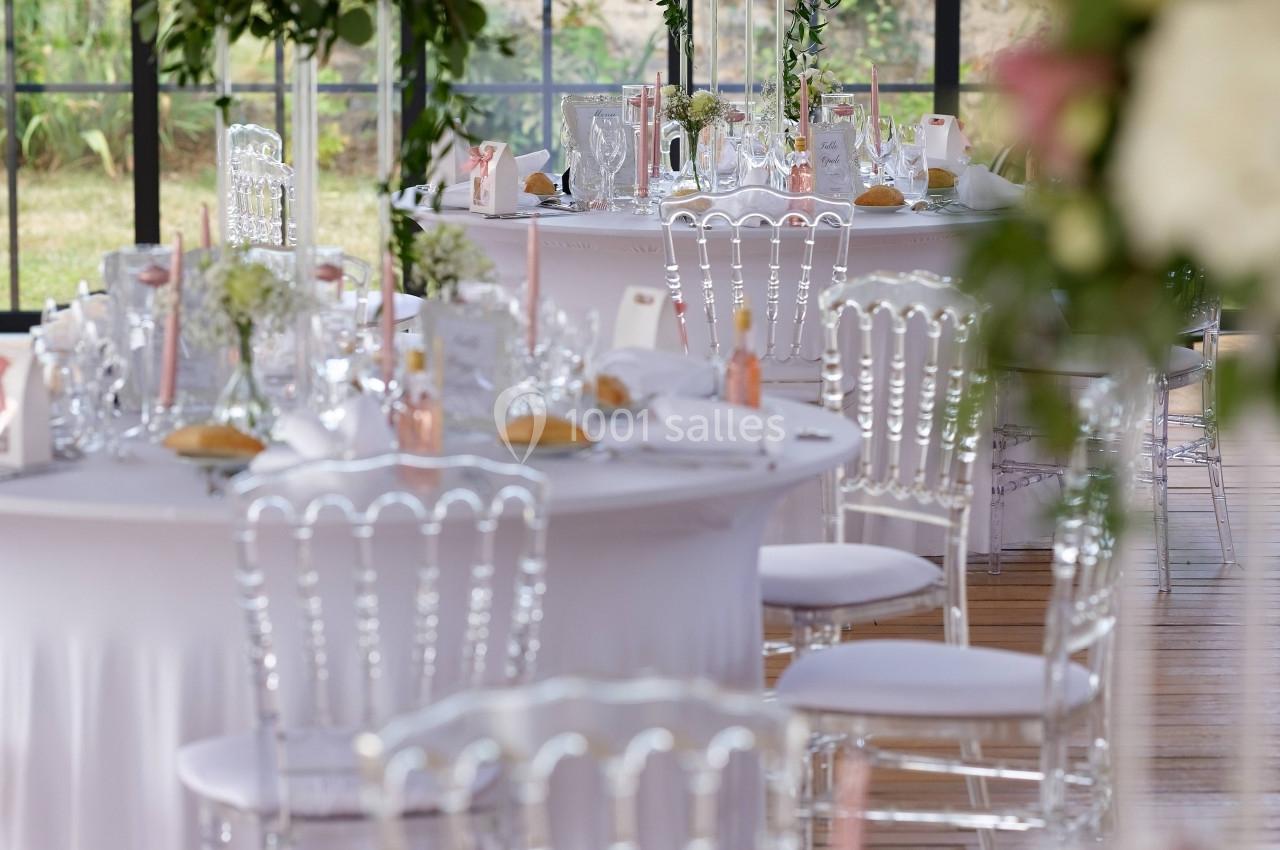 Salle de réception décorée avec des tables rondes, nappes blanches, chaises transparentes et centres de table fleuris.