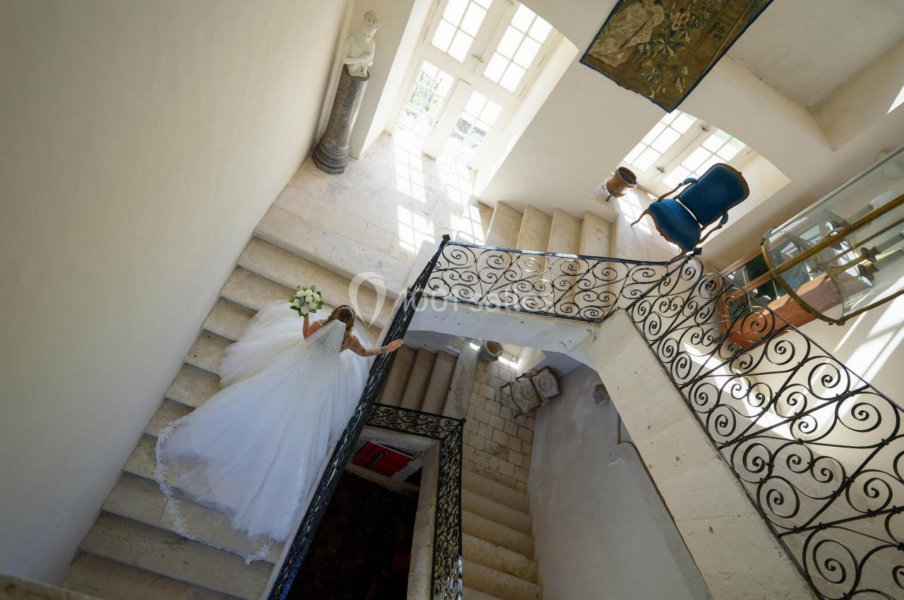 Une mariée en robe blanche descend un escalier en pierre dans un intérieur lumineux avec des rampes en fer forgé.