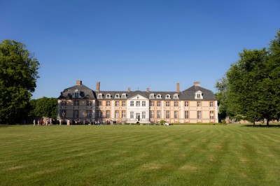 Façade d'un château en pierre et briques entouré d'une pelouse et d'arbres sous un ciel bleu clair.