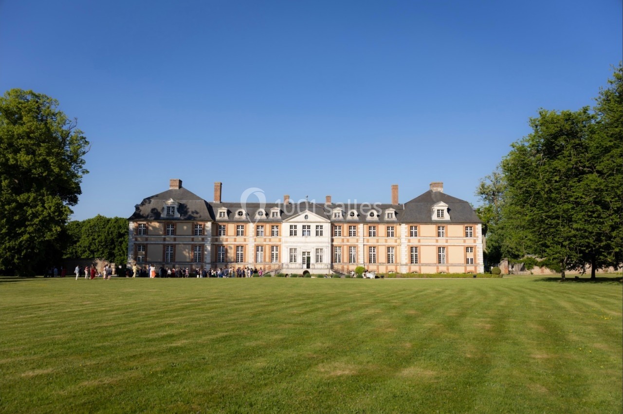 Façade d'un château en pierre et briques entouré d'une pelouse et d'arbres sous un ciel bleu clair.