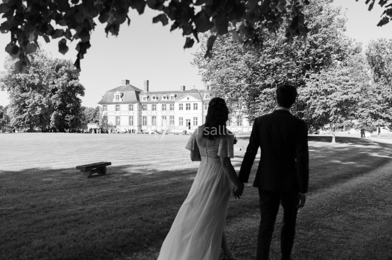 Un couple marche main dans la main vers un grand bâtiment historique entouré d'arbres, par une journée ensoleillée.