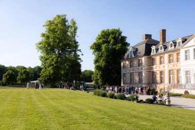 Façade d'un château en pierre et briques entouré d'une pelouse et d'arbres sous un ciel bleu clair.