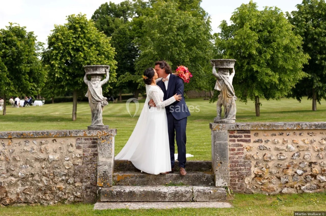 Un couple en tenue de mariage s'embrasse sur un escalier en pierre, entouré de statues et d'un parc verdoyant.