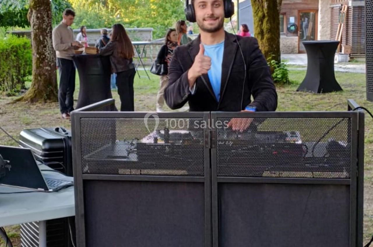 Un DJ souriant derrière une table de mixage en extérieur, avec des invités discutant en arrière-plan.