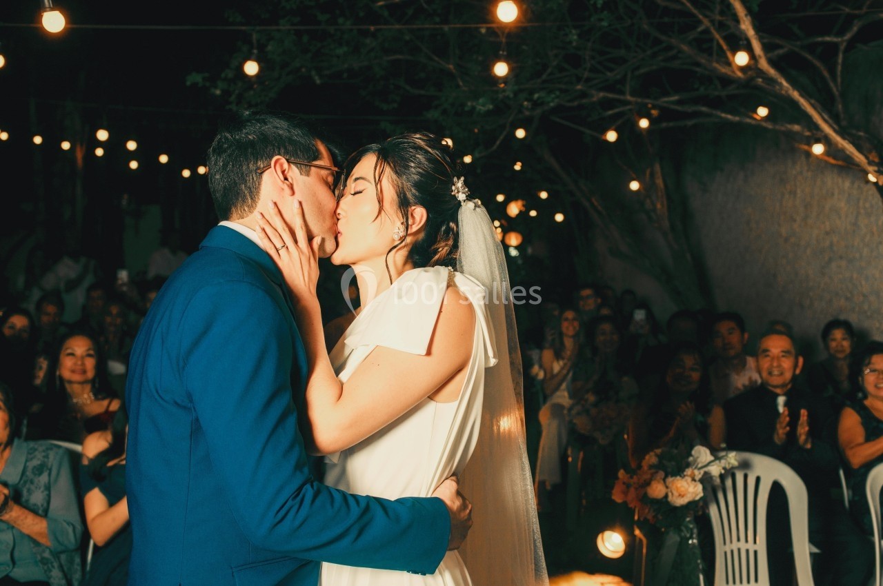 Un couple s'embrasse lors de leur mariage en soirée, entouré d'invités et de guirlandes lumineuses.