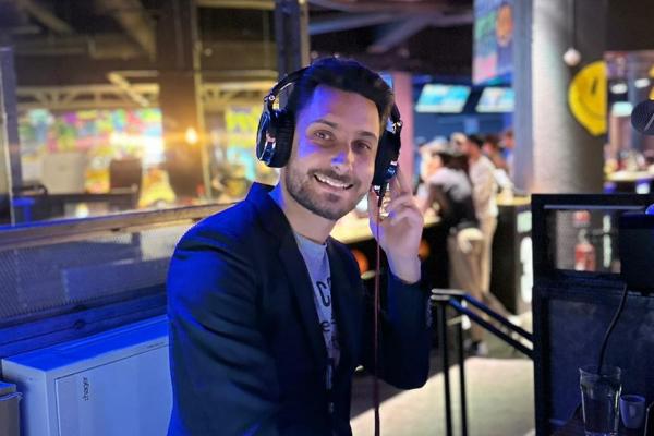 Homme souriant portant un casque audio, debout dans un environnement lumineux avec des écrans et des personnes en arrière…