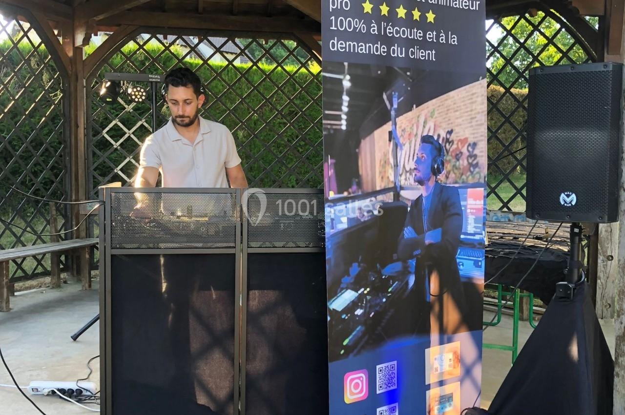 Un DJ mixe sous un kiosque en bois, avec une bannière promotionnelle et du matériel audio installé devant lui.