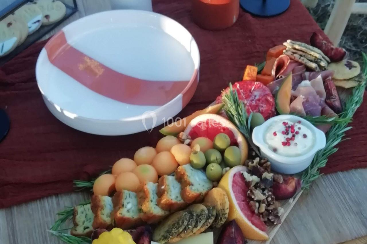 Plateau de charcuterie et fruits avec fromages, dips et garnitures sur une table en bois, décorée pour un repas en extérieur.