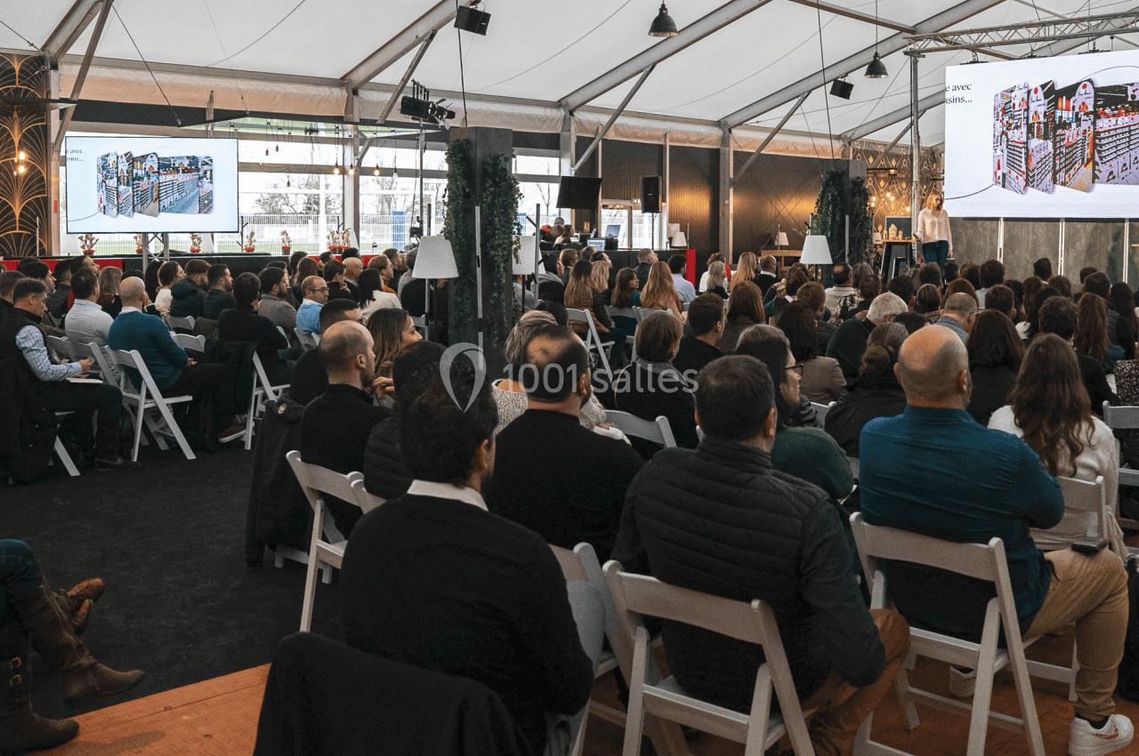 Un public assis dans une salle sous tente assiste à une présentation projetée sur un écran géant.