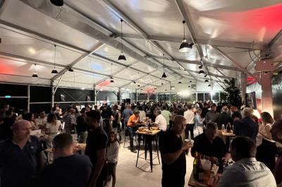 Groupe de personnes rassemblées sous une tente lors d'un événement en extérieur, près d'un bâtiment moderne.