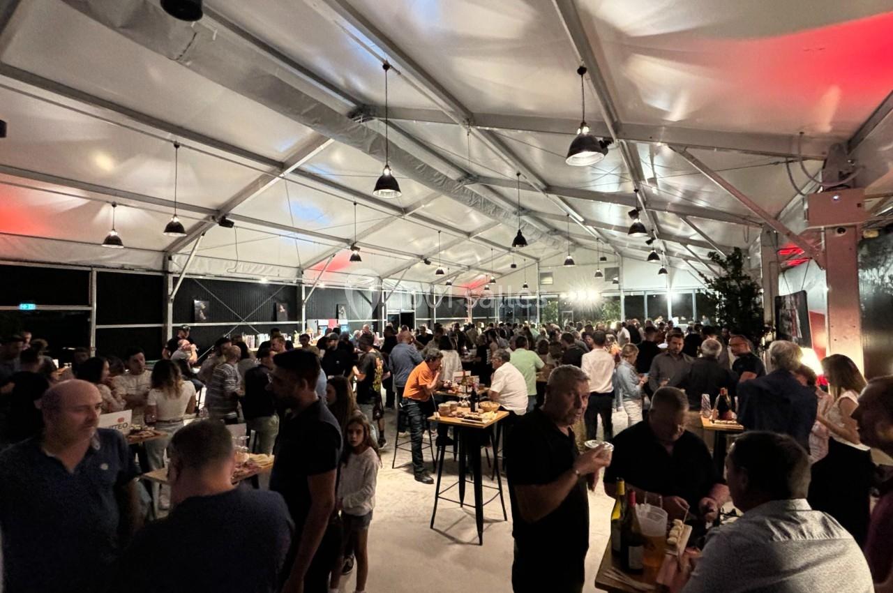 Personnes rassemblées dans une grande tente éclairée, discutant et partageant un moment convivial autour de tables hautes.