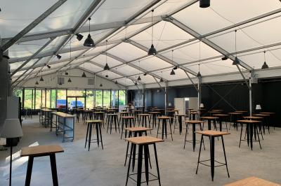 Groupe de personnes rassemblées sous une tente lors d'un événement en extérieur, près d'un bâtiment moderne.