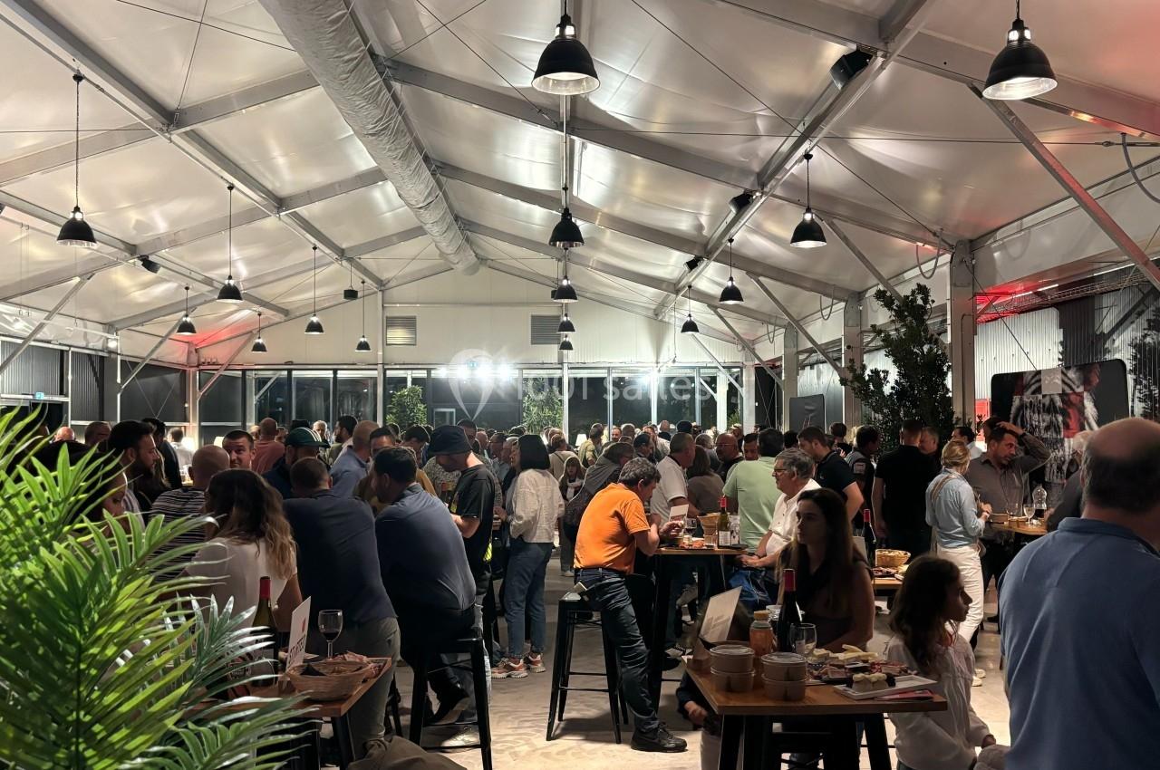 Salle lumineuse sous une structure en toile, remplie de personnes discutant autour de tables lors d'un événement.