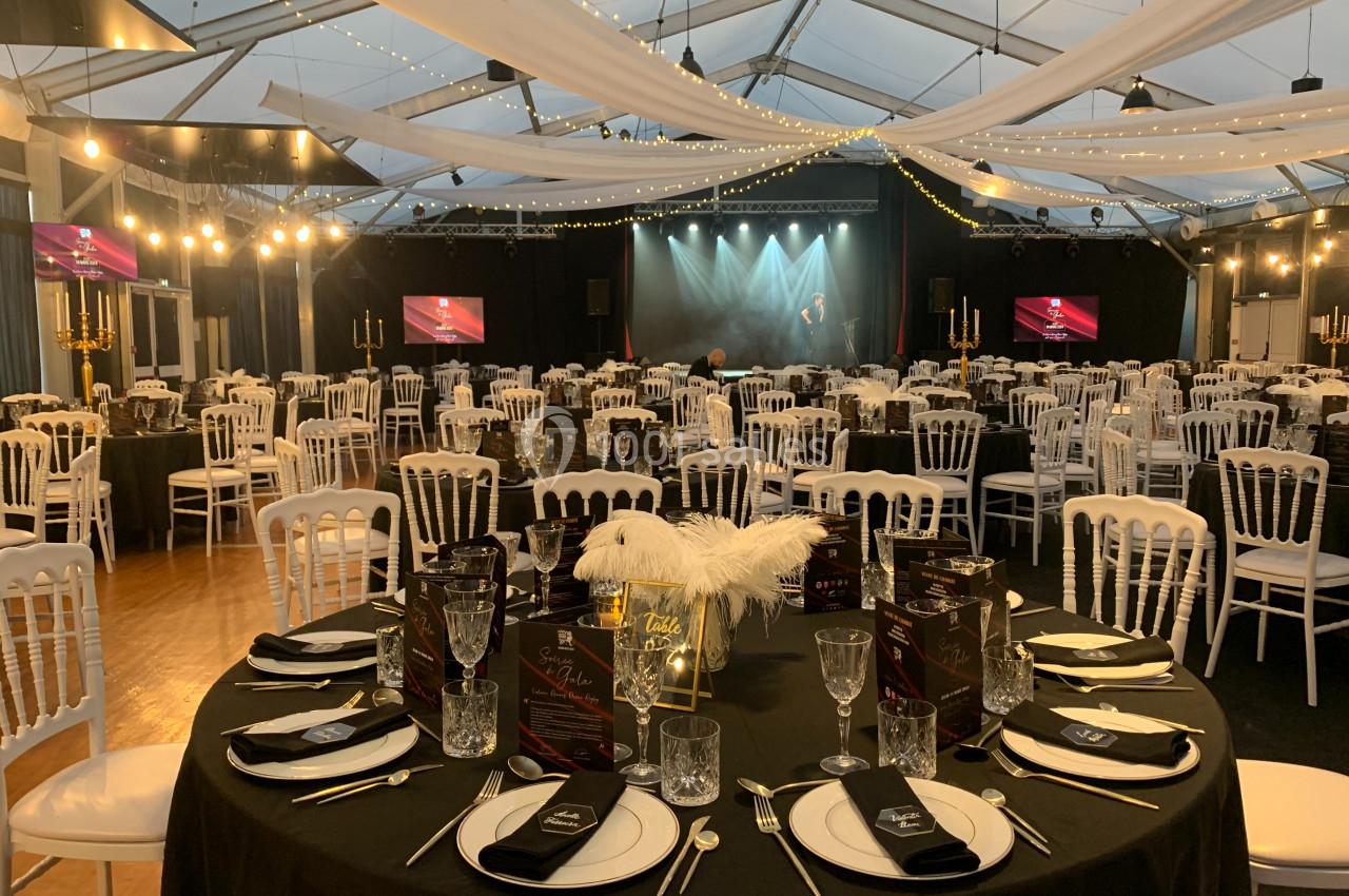 Salle de réception décorée avec des tables rondes dressées, nappes noires, chaises blanches et éclairage chaleureux.