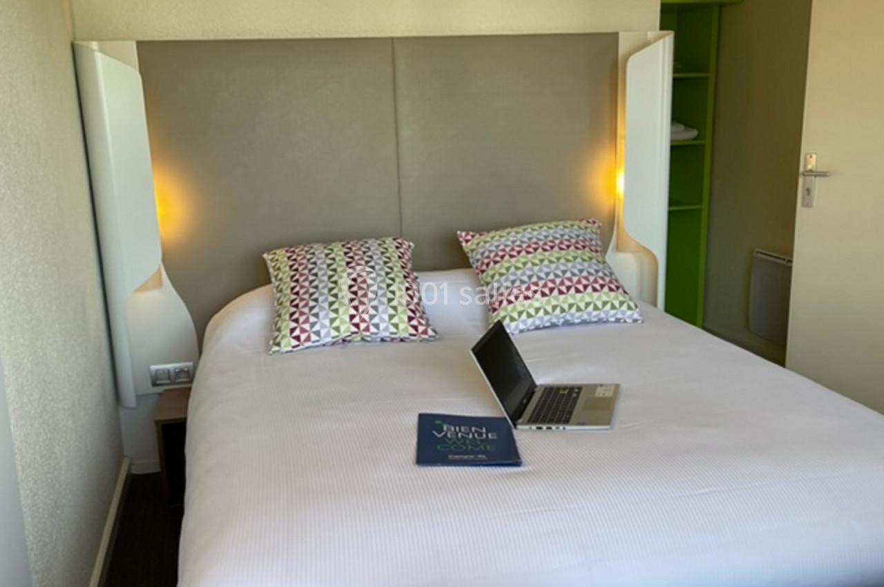 Lit double avec draps blancs, deux coussins à motifs colorés, un ordinateur portable et un livret posés dessus.