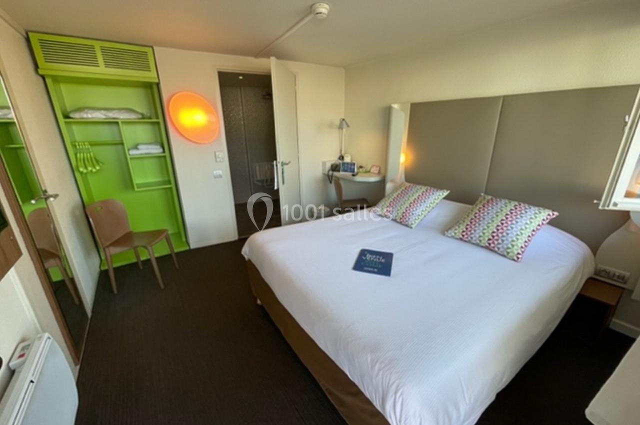 Chambre d'hôtel avec lit double, tête de lit rembourrée, étagères vertes ouvertes et salle de bain visible en arrière-plan.