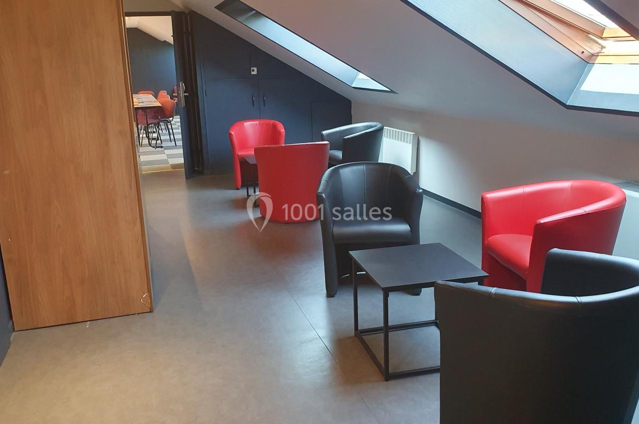 Espace mansardé avec fauteuils rouges et noirs autour de petites tables, éclairé par des fenêtres de toit.