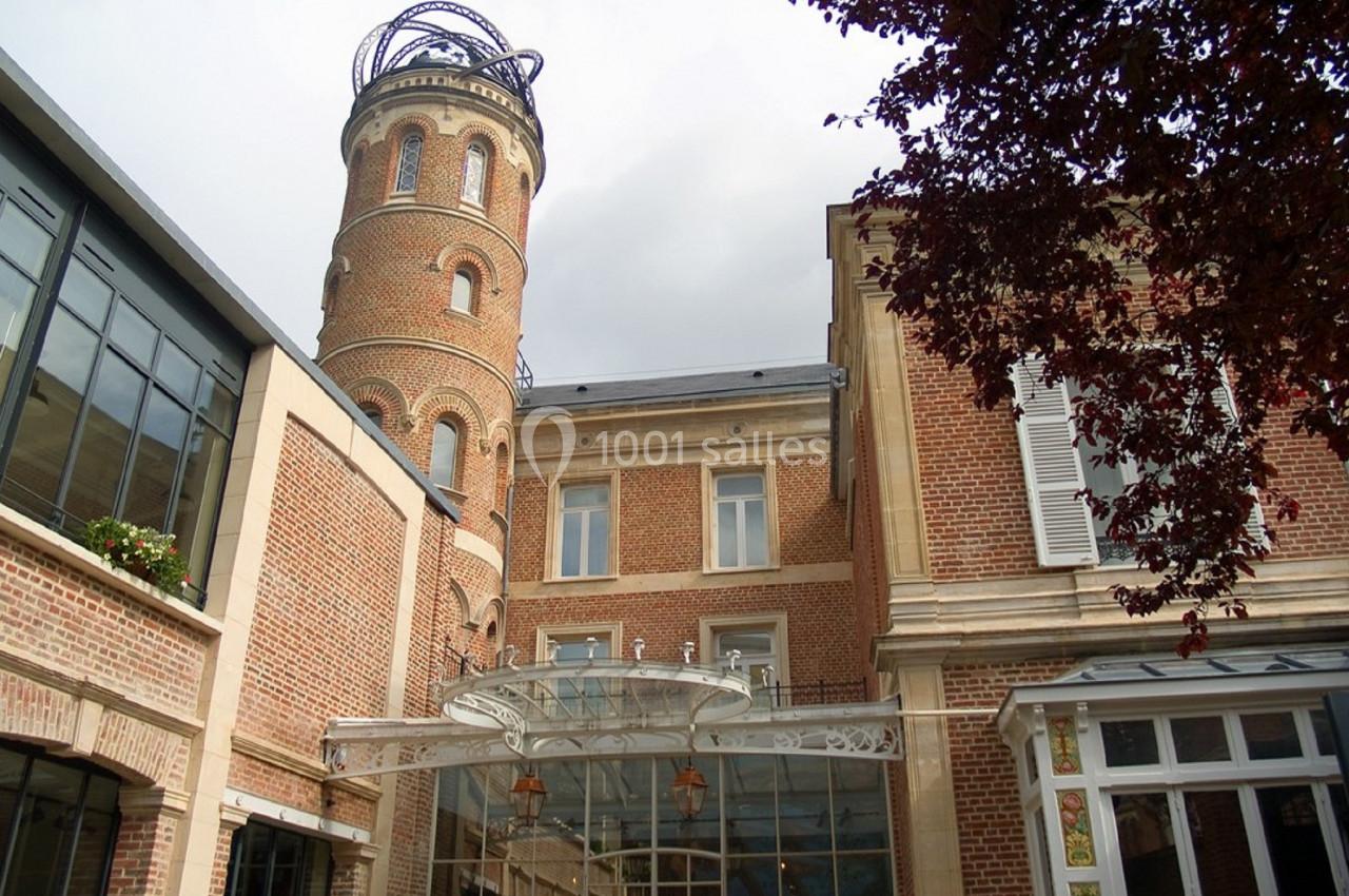 Bâtiment en briques rouges avec une tour cylindrique et une verrière, entouré d'arbres et d'éléments modernes.