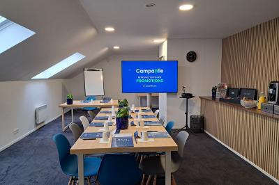 Salle de réunion lumineuse avec table rectangulaire, chaises colorées, écran, paperboard et coin café en arrière-plan.