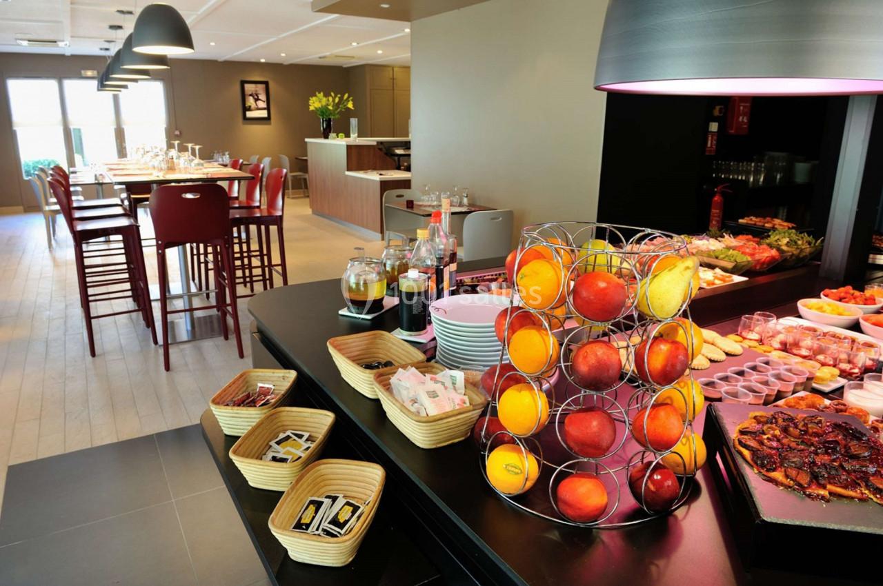 Buffet de petit-déjeuner avec fruits frais, viennoiseries, boissons et condiments dans une salle lumineuse.