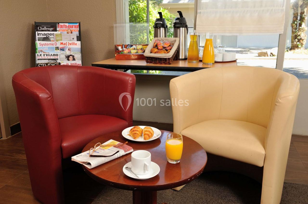 Deux fauteuils autour d'une table avec croissants, jus d'orange et café, près d'un buffet et de magazines.