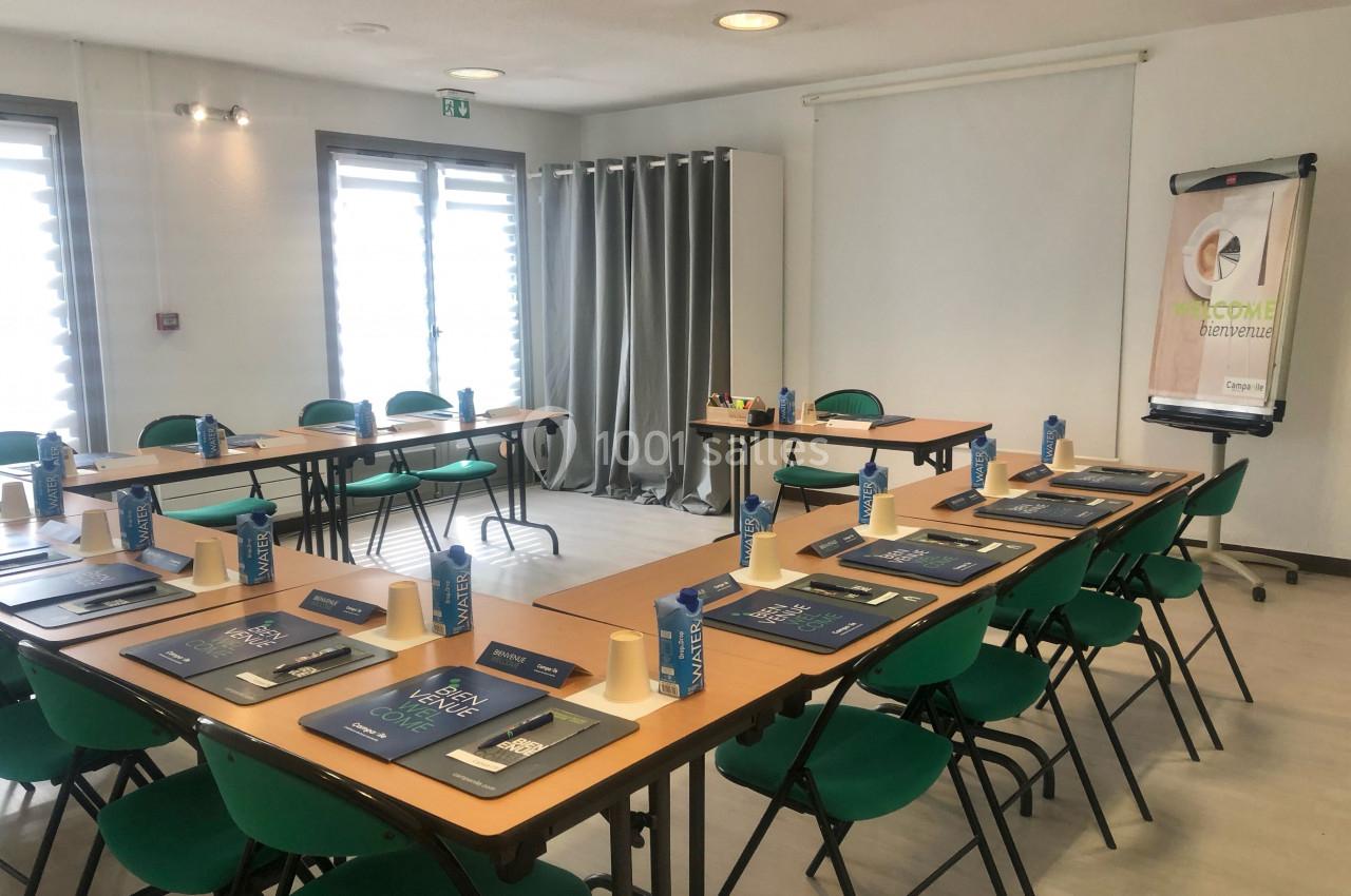 Salle de réunion lumineuse avec tables disposées en U, chaises vertes et matériel de conférence installé.