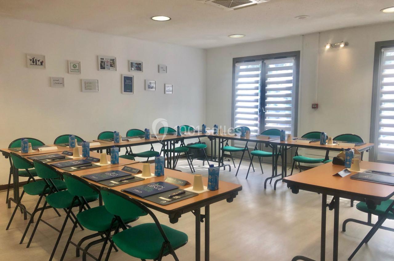 Salle de réunion lumineuse avec tables disposées en U, chaises vertes et matériel de travail sur chaque place.