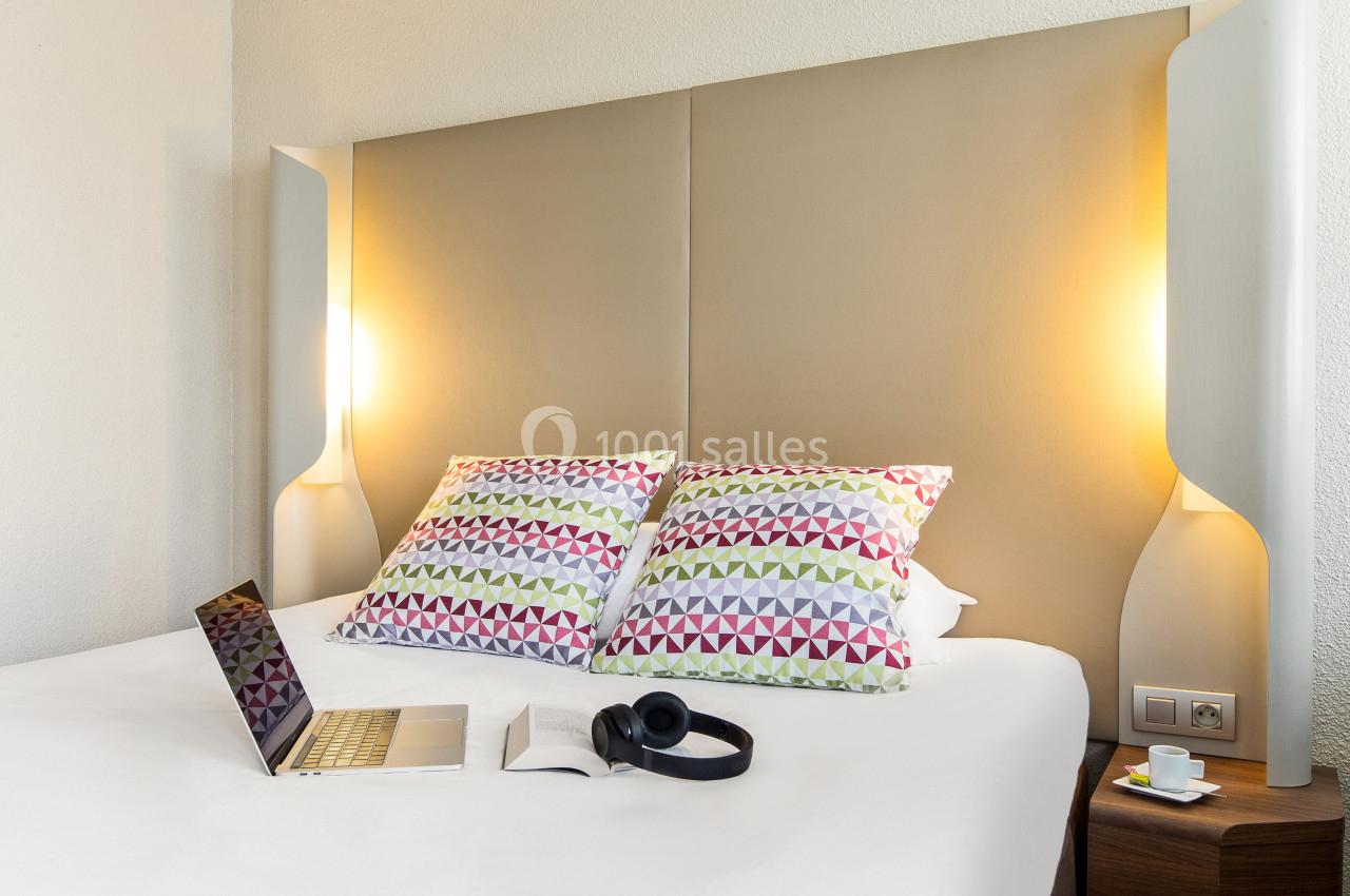 Lit double avec tête de lit beige, deux coussins colorés, un ordinateur portable et un casque audio posés dessus.