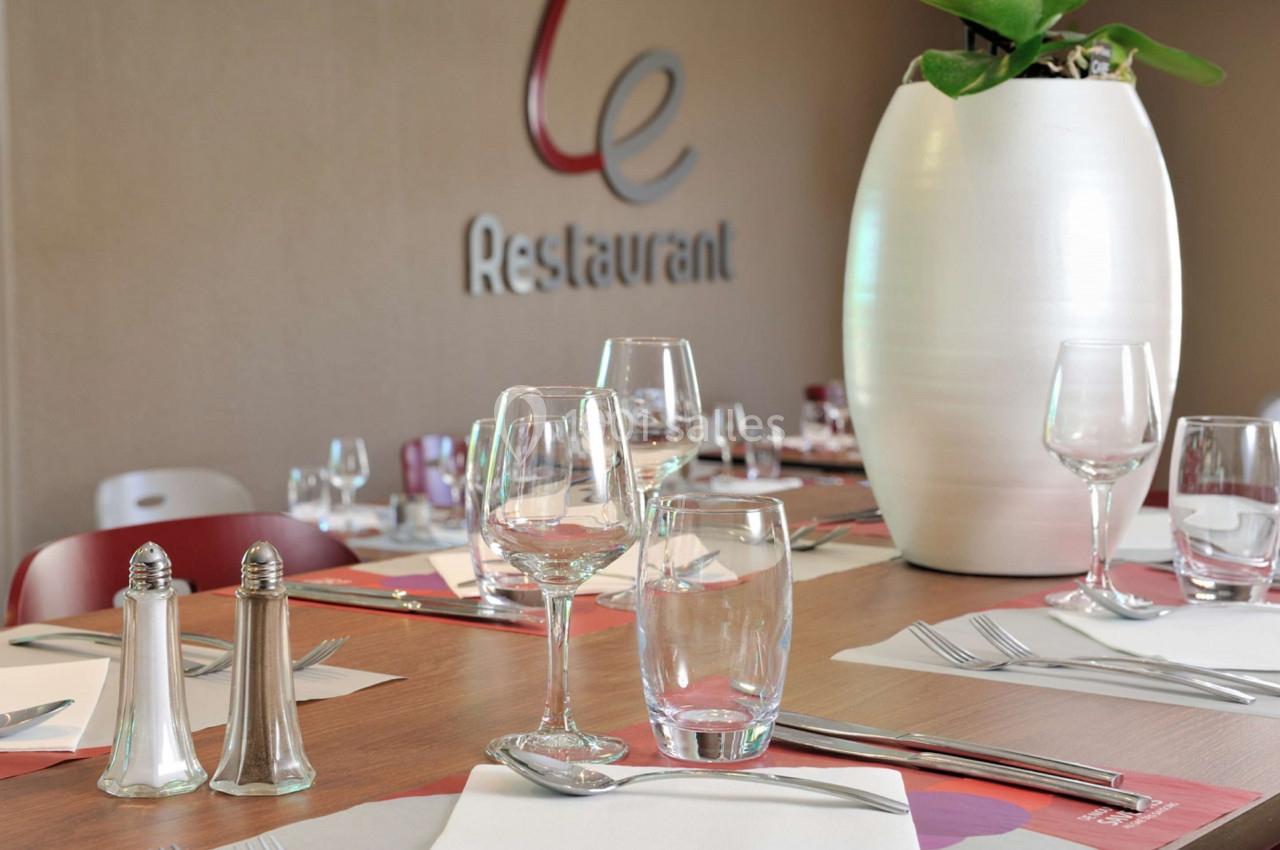 Table dressée avec verres, couverts et serviettes, devant un mur beige avec un logo et une plante en pot blanc.